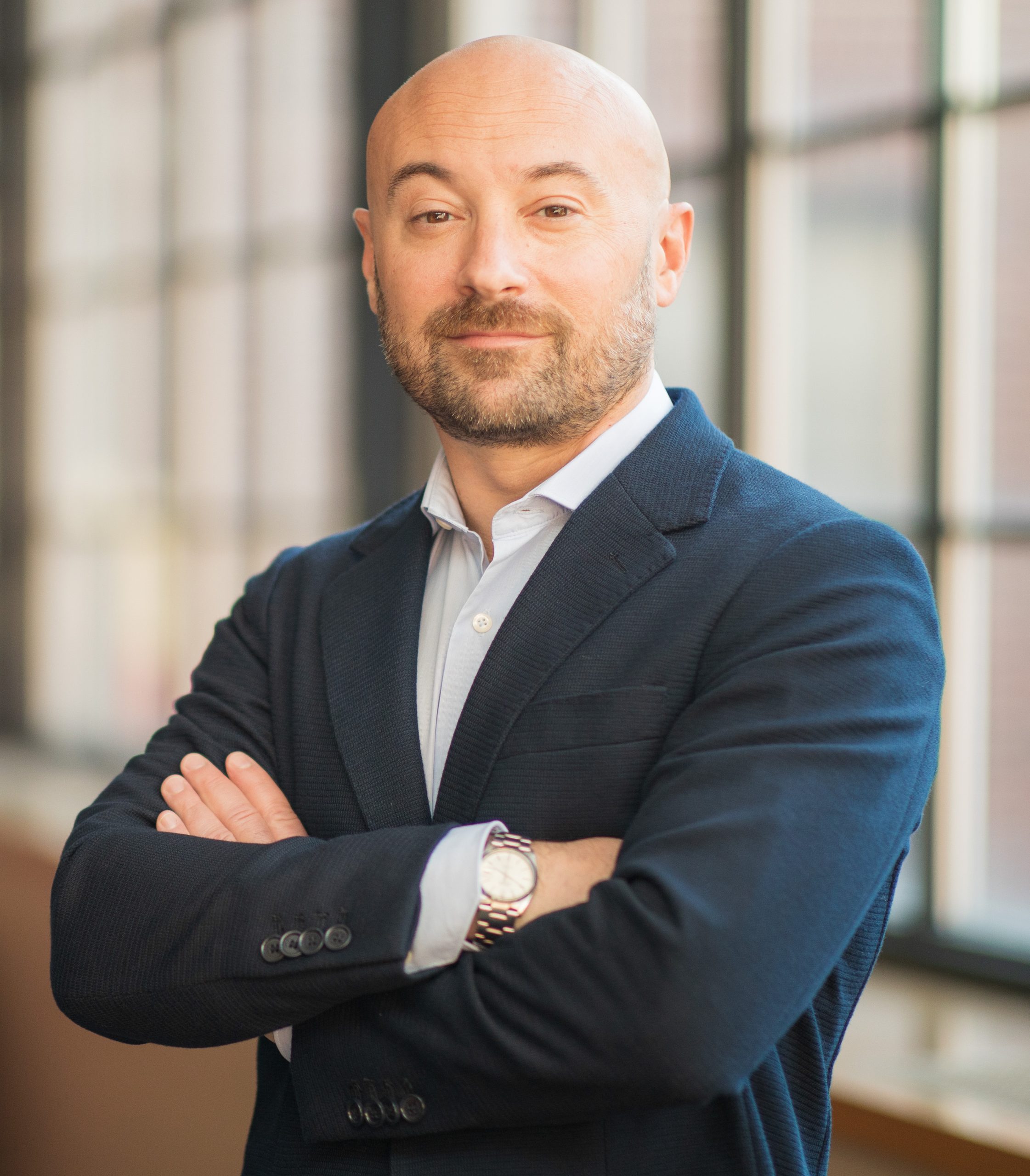 A professional headshot of a man in a blue suit jacket, light button up shirt, standing with his arms crossed in front of windows